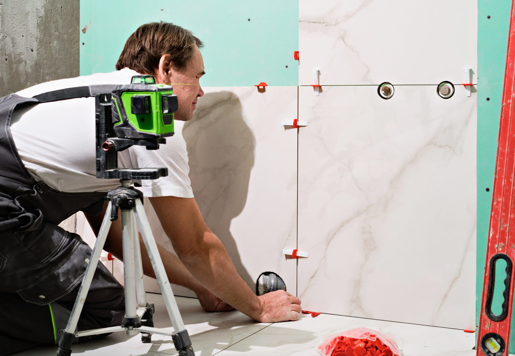 Professional master man laying ceramic tiles on wall in bathroom. Portrait of an experienced repairman laying large size porcelain tiles.Construction worker checking tile installation with laser level