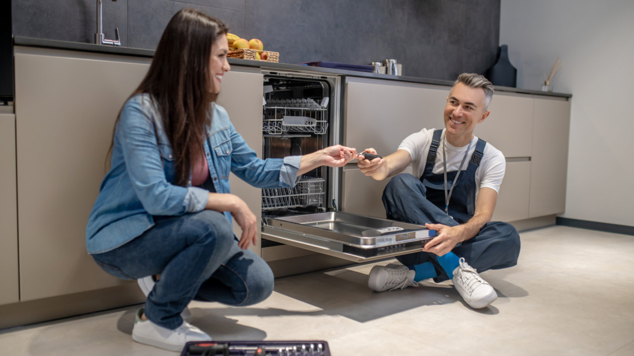 Household repair. Smiling long-haired middle-aged woman crouching holding out tool to joyful plumber sitting on floor near open dishwasher in kitchen