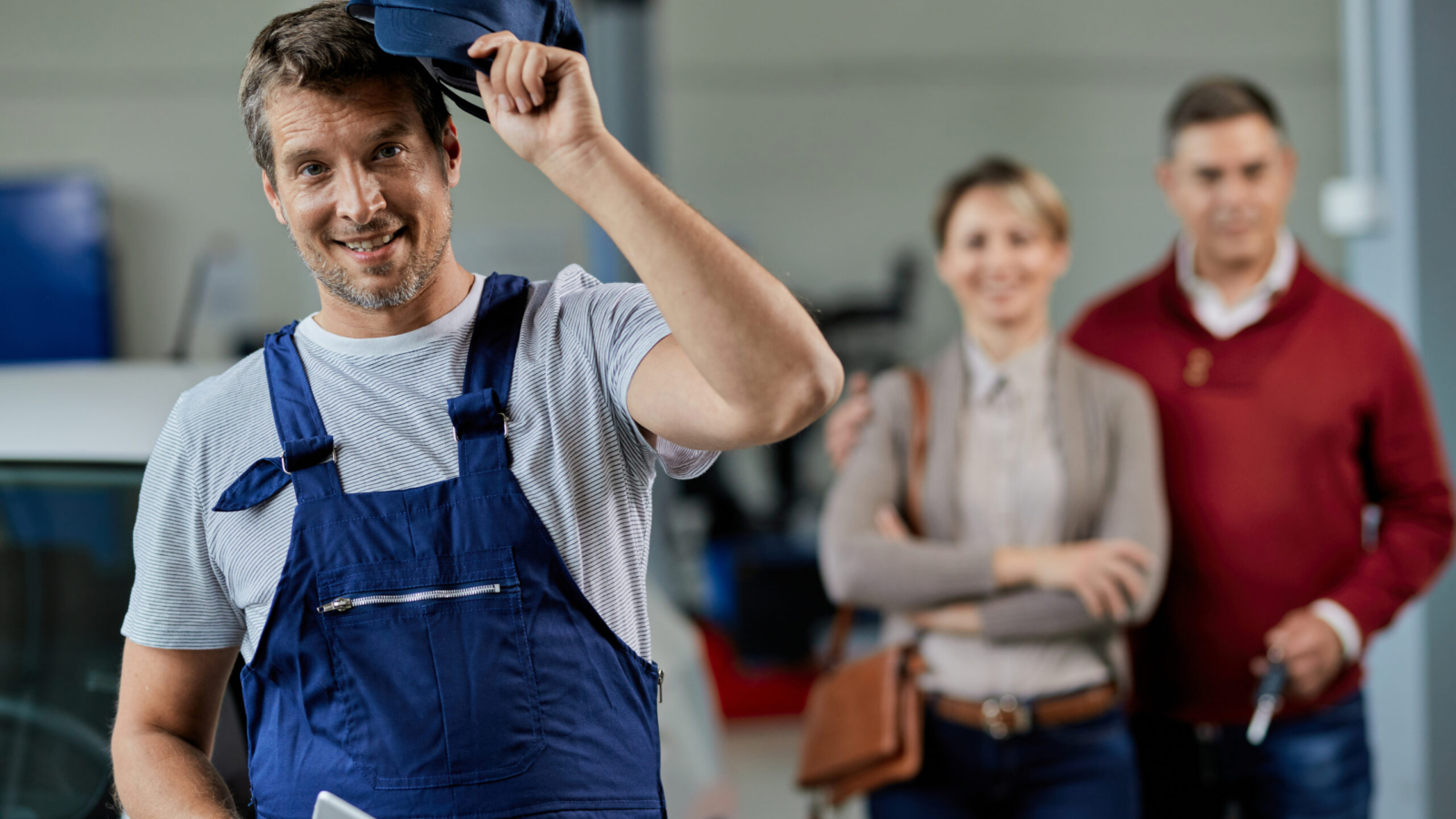Happy auto mechanic taking his hat of while standing in a workshop and looking at camera. His customers are in the background.