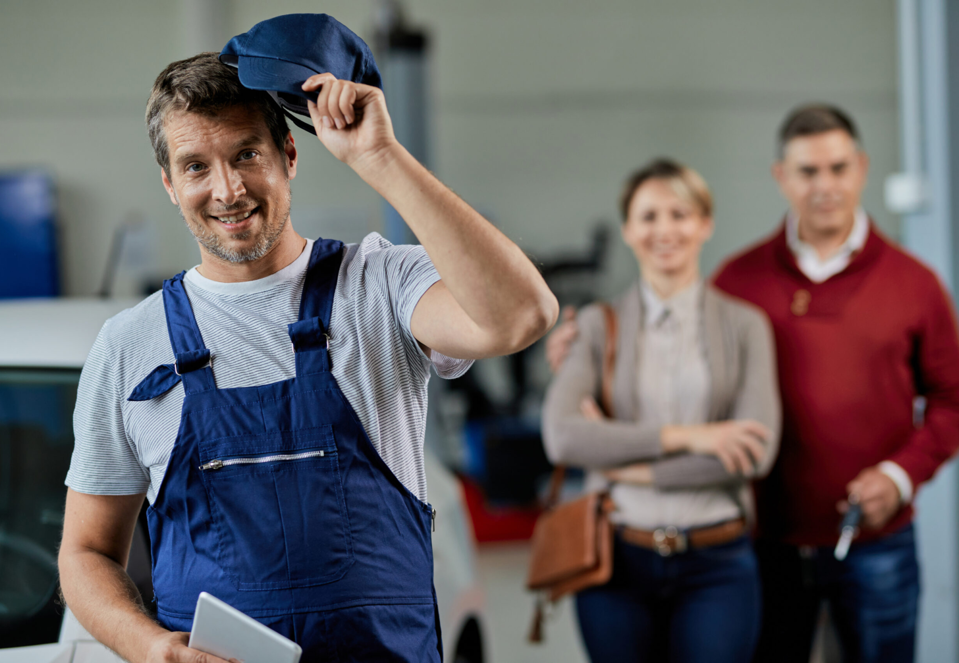 Happy auto mechanic taking his hat of while standing in a workshop and looking at camera. His customers are in the background.